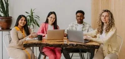Diverse group of four professionals smiling and working on laptops at a wooden table, reflecting BitBop's growing team.