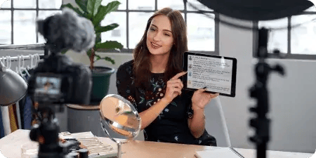 Woman sitting at a desk with a makeup setup, recording content in front of a camera while holding a tablet showing social media insights — representing a Social Media Manager role at BitBop in Islamabad, Pakistan.