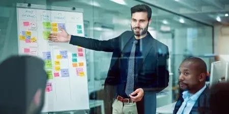 Professional team in a modern office discussing strategy as a man presents colorful post-it notes on a glass board.
