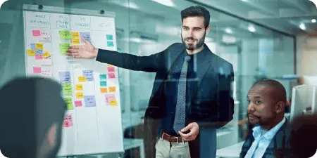 A marketing professional in a formal setting presenting PPC and ad strategy on a whiteboard covered with sticky notes, addressing a team in a modern office environment.