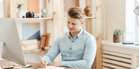 Young man smiling while writing at a desk with a desktop computer, in a bright office setting, applying for a content role.